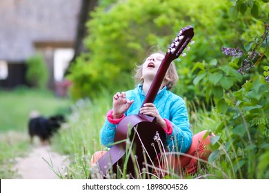 Little Girl Playing Guitar And Singing Outdoors On Green Meadow At Spring. Happy Child With Musical Instrument On Rural Yard.