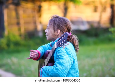 Little Girl Playing Guitar And Singing Outdoors On Green Meadow At Spring. Happy Child With Musical Instrument On Rural Yard.