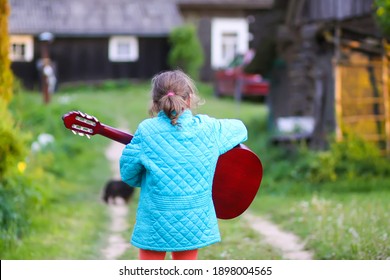 Little Girl Playing Guitar And Singing Outdoors On Green Meadow At Spring. Happy Child With Musical Instrument On Rural Yard.