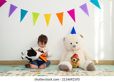 Little Girl Playing With Her Soft Toys Imagining A Birthday Party. She Is Putting On A Cone Hat To A Cow Soft Toy. 