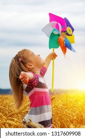 Little Girl Playing On The Meadow On Sun With Windmill In Her Hands.  Child Holding Wind Toy  On Wheat Field.