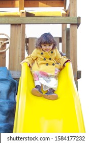 Little Girl Playing On Yellow Slide Wearing A Yellow Trench Coat. She Has Brown Curly Hair And Is On A Child's Play Set At Day Care.