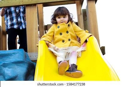 Little Girl Playing On Yellow Slide Wearing A Yellow Trench Coat. She Has Brown Curly Hair And Is On A Child's Play Set At Day Care.