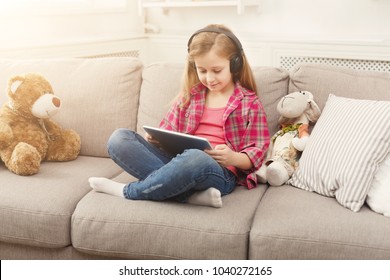 Little Girl Playing Online Games On Tablet And Listening To Music In Headphones. Female Child Sitting On Sofa With Her Teddy Bear. Kid Leisure At Home. Social Networking And Online Education Concept