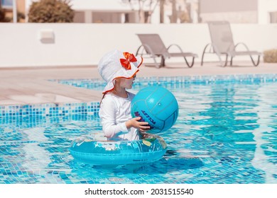 Little Girl Playing In Pool With Ball In Swimwear With Sun Protection. Healthy And Safe Summer Outdoor Activities. Summer Tropical Vacation