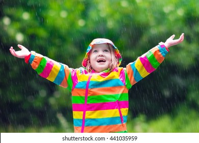 Little girl playing in rainy summer park. Child with rainbow umbrella, waterproof coat and boots jumping in puddle in the rain. Kid walking in autumn shower. Outdoor fun by any weather