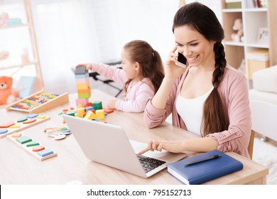 A Little Girl Is Playing At The Table While Her Mother Is Talking On The Phone. It's Mom And Daughter. On The Table Is Also A Laptop.