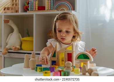 A Little Girl Playing With Wooden Blocks On The Table In Playroom.  Educational Game For Baby And Toddler In Modern Nursery. The Kid Builds A Tower From Wooden Rainbow Stacking Blocks.