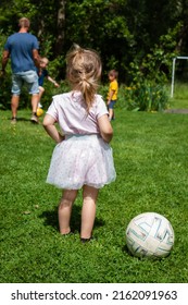 A Little Girl With A Ponytail, A Pink Dress And Tutu Standing Next To A Soccer Ball With Her Hands On Hips. A Group Of Kids Playing Football In The Garden In The Background With A Grownup Man.