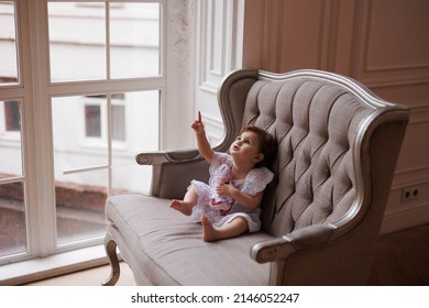 Little Girl Portrait. Small Baby Kid Playing With A Doll. Serious Little Daughter Looking Attentively And Thoughtfully. One-year-old Girl In Whire Dress Sitting On Beige Sofa In Big Room. Sweet Kid