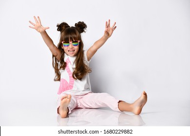 Little Girl Posing Sitting On The Floor In Dark Glasses, Summer Clothes And Fashionable Hairstyles, In The Studio, Arms And Legs Stretched Out In Different Directions