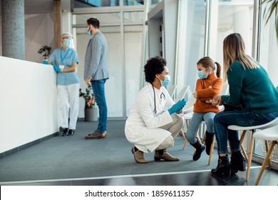 Little Girl Pouting While African American Female Doctor Is Talking To Her In A Waiting Room At The Hospital. They Are Wearing Protective Face Mask Dur To Coronavirus Epidemic. 