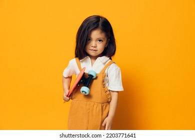 A Little Girl Of Preschool Age Stands On An Orange Background In An Orange Dress And A White Shirt Holding A Skate In Her Hands, Looking Menacingly At The Camera