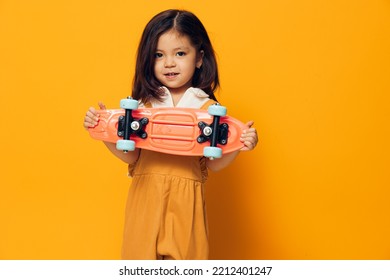 A Little Girl Of Preschool Age Stands On An Orange Background In An Orange Dress And A White Shirt Holding A Skate In Front Of Her, Smiling Looking At The Camera