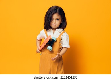 A Little Girl Of Preschool Age Stands On An Orange Background In An Orange Dress And A White Shirt Holding A Skate In Her Hands, Looking Menacingly At The Camera