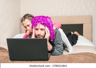 A Little Girl Prevents Dad From Working At A Computer. A Child Puts A Bright Wig On The Head Of A Bald Man.telecommuting In Quarantine. A Fun Family Event.