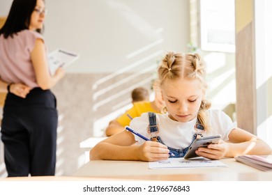 Little Girl Primary School Girl Student With Two Braids In Classroom Cheating With Smartphone During Classwork At Classroom. Attendancy, Academic Performance Concept.