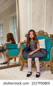 Little Girl Princess 5-6 Years Old, Fashion Model In Stylish Elegant Blue Dress Sitting On Armchair In Living Room With Mirror, Looking At Camera. Fashionable Young Lady Actress At Home. Copy Space