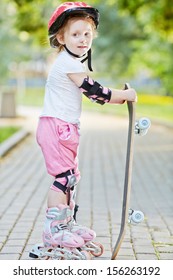 Little girl in protective equipment and rollers stands leaning on skateboard in park.