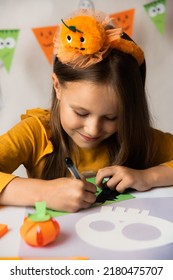 A Little Girl In A Pumpkin Costume Draws A Mask With A Black Felt-tip Pen. Halloween Party. Craft For Kid