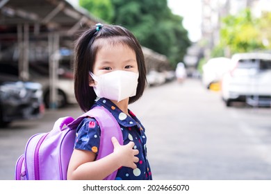 Little Girl With A Purple Backpack Over Her Shoulders Turn Around With A Smile On Her Face. Concept Back To School. Kid Wearing 3D Surgical Face Mask. Child Aged 4-5 Years Old.