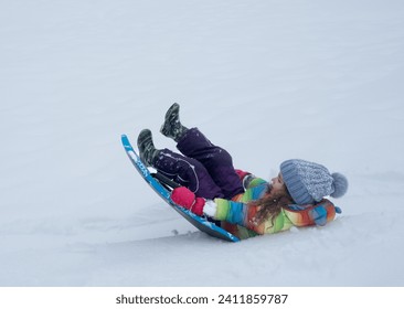 Little girl with rainbow jacket sledging down a snowy hill, falling in a snow. - Powered by Shutterstock