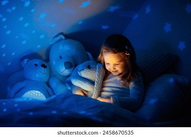 Little girl reading a book in bed. Dark bedroom with night light projecting stars on room ceiling. Kids nursery and bedding. Children read before bedtime. Toddler child playing with lamp and bear toy. - Powered by Shutterstock