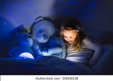 Little Girl Reading A Book In Bed. Dark Bedroom With Night Light Projecting Stars On Room Ceiling. Kids Nursery And Bedding. Children Read Before Bedtime. Toddler Child Playing With Lamp And Bear Toy.