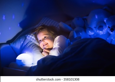 Little Girl Reading A Book In Bed. Dark Bedroom With Night Light Projecting Stars On Room Ceiling. Kids Nursery And Bedding. Children Read Before Bedtime. Toddler Child Playing With Lamp And Bear Toy.