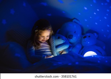 Little Girl Reading A Book In Bed. Dark Bedroom With Night Light Projecting Stars On Room Ceiling. Kids Nursery And Bedding. Children Read Before Bedtime. Toddler Child Playing With Lamp And Bear Toy.