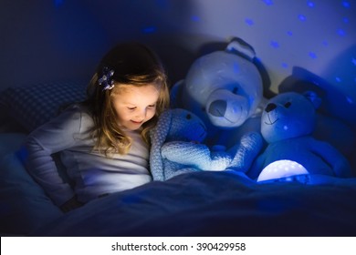 Little Girl Reading A Book In Bed. Dark Bedroom With Night Light Projecting Stars On Room Ceiling. Kids Nursery And Bedding. Children Read Before Bedtime. Toddler Child Playing With Lamp And Bear Toy.