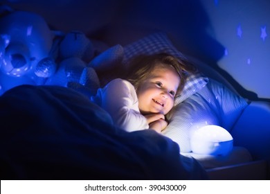 Little Girl Reading A Book In Bed. Dark Bedroom With Night Light Projecting Stars On Room Ceiling. Kids Nursery And Bedding. Children Read Before Bedtime. Toddler Child Playing With Lamp And Bear Toy.