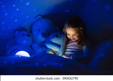 Little Girl Reading A Book In Bed. Dark Bedroom With Night Light Projecting Stars On Room Ceiling. Kids Nursery And Bedding. Children Read Before Bedtime. Toddler Child Playing With Lamp And Bear Toy.