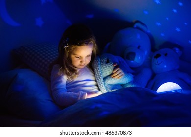 Little Girl Reading A Book In Bed. Dark Bedroom With Night Light Projecting Stars On Room Ceiling. Kids Nursery And Bedding. Children Read Before Bedtime. Toddler Child Playing With Lamp And Bear Toy.