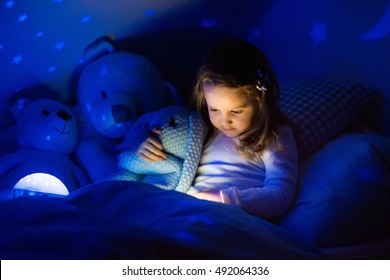 Little Girl Reading A Book In Bed. Dark Bedroom With Night Light Projecting Stars On Room Ceiling. Kids Nursery And Bedding. Children Read Before Bedtime. Toddler Child Playing With Lamp And Bear Toy.