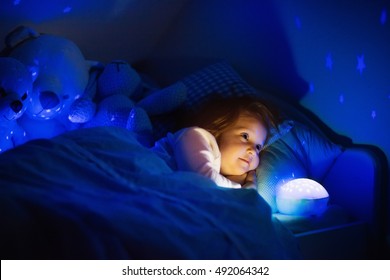 Little Girl Reading A Book In Bed. Dark Bedroom With Night Light Projecting Stars On Room Ceiling. Kids Nursery And Bedding. Children Read Before Bedtime. Toddler Child Playing With Lamp And Bear Toy.