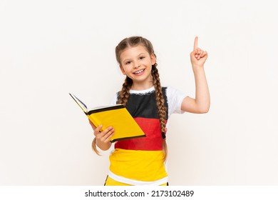 A little girl is reading a book in German and pointing up at your advertisement. Courses for teaching German to children. A child in a jacket with a German flag on a white isolated background.