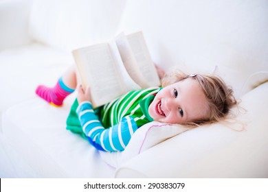 Little Girl Reading A Book Relaxing On A White Couch. Kids Read Books At Home Or Preschool. Children Learning And Doing Homework After School. Child Playing. Toddler Kid In Colorful Dress On A Sofa.