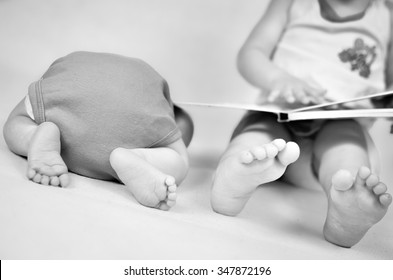 Little Girl Is Reading A Book Tog Her Baby Brother. Black And White Photo With Soft Focus On Their Feet. Family Values