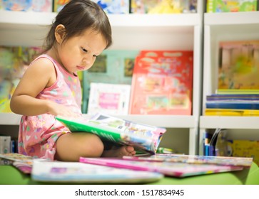 Little Girl Reading Next To A Stack Of Books In The  Library.