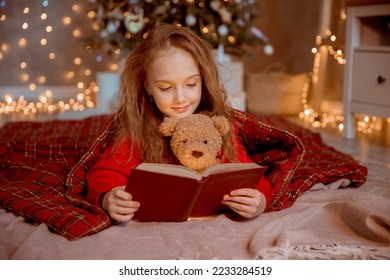 a little girl reads a book with a teddy bear near the Christmas tree at home for Christmas - Powered by Shutterstock