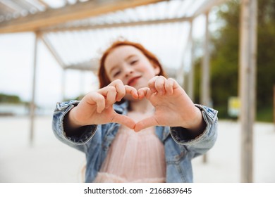 A Little Girl With Red Hair Smiles And Shows A Heart Sign With Her Hands. A Child On The Beach.