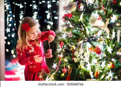 Little Girl In Red Knitted Nordic Reindeer Sweater Hanging Ornaments On Christmas Tree With Light, Bauble And Candy Canes. Child Decorating Xmas Tree In Beautiful Family Living Room With Fireplace.