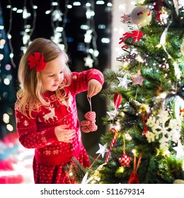 Little Girl In Red Knitted Nordic Reindeer Sweater Hanging Ornaments On Christmas Tree With Light, Bauble And Candy Canes. Child Decorating Xmas Tree In Beautiful Family Living Room With Fireplace.