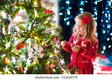 Little Girl In Red Knitted Nordic Reindeer Sweater Hanging Ornaments On Christmas Tree With Light, Bauble And Candy Canes. Child Decorating Xmas Tree In Beautiful Family Living Room With Fireplace.