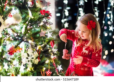 Little girl in red knitted Nordic reindeer sweater hanging ornaments on Christmas tree with light, bauble and candy canes. Child decorating Xmas tree in beautiful family living room with fireplace.
