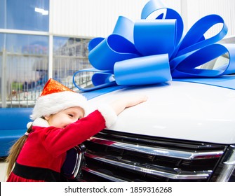 Little girl in red Santa Claus outfit hugs the car in front. Auto as a present with a big blue bow. The child's face is not visible
