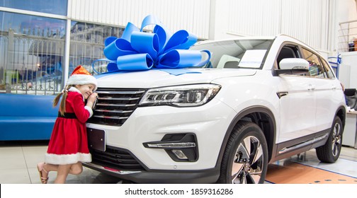 Little girl in red Santa Claus outfit hugs the car in front. Auto as a present with a big blue bow. 