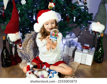 A little girl in a red Santa's Hat is sitting under the Christmas tree and playing with a magic snow globe. Christmas and New Year atmosphere. Concept of a happy childhood. Slow motion. - Powered by Shutterstock