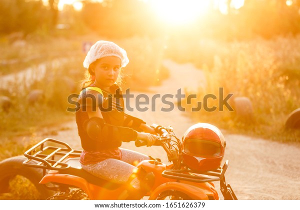 The little\
girl rides a quad bike ATV. A mini quad bike is a cool girl in a\
helmet and protective clothing. Electric quad bike electric car for\
children popularizes green\
technology.